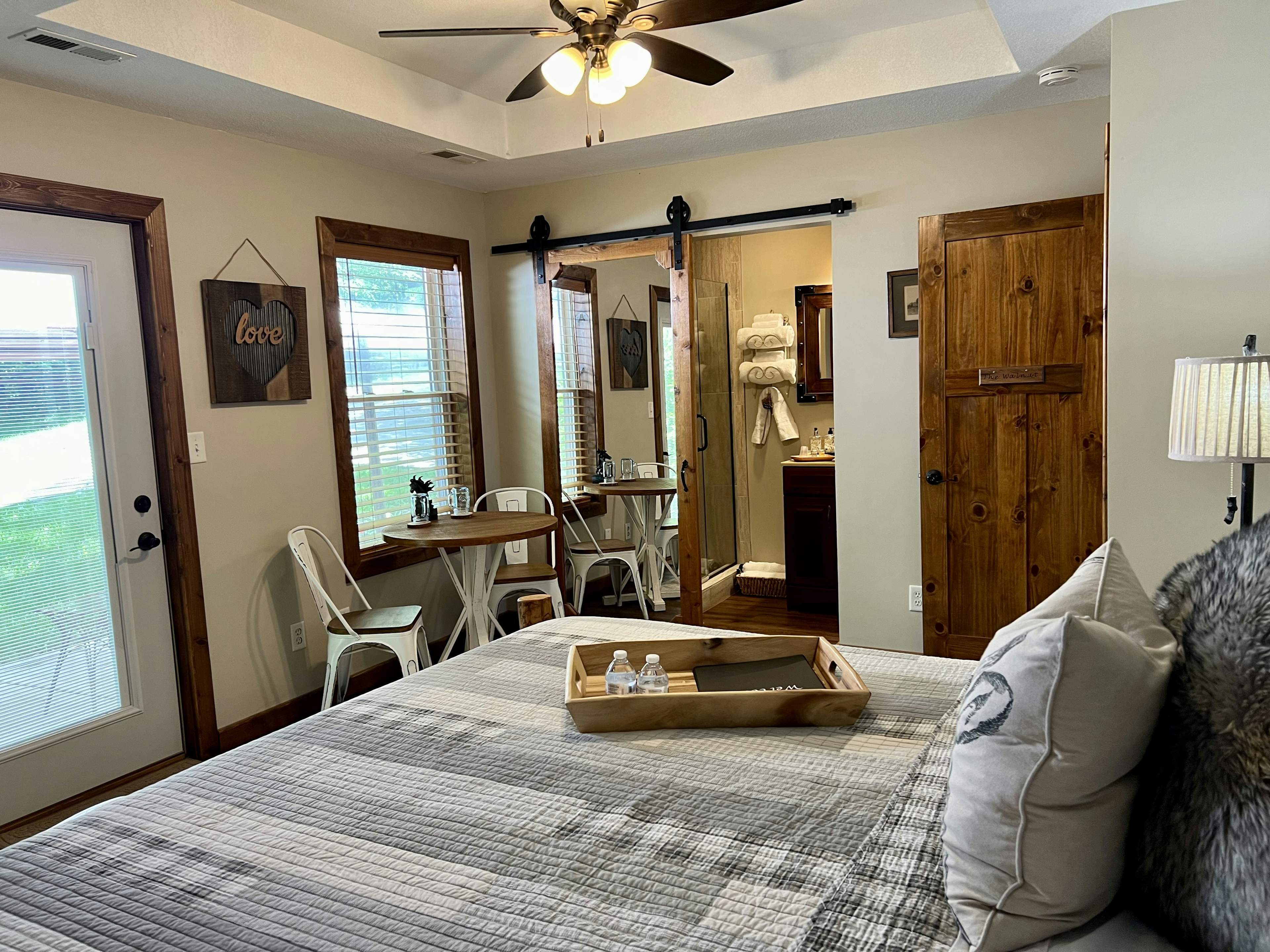 log style bed with white and grey country quilt, single door to outside, one small window, mirrored barn style door open to bathroom, ceiling fan