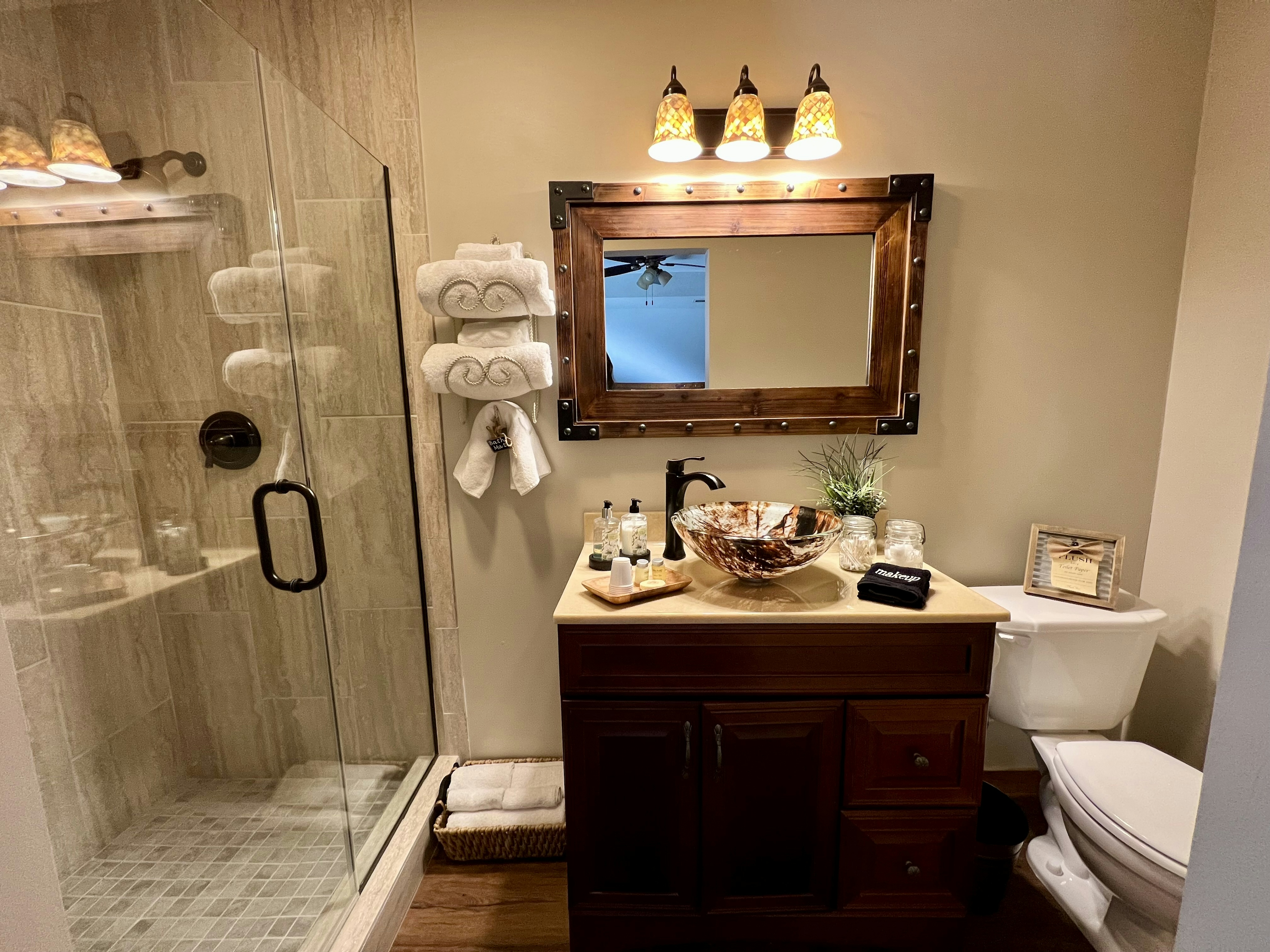 bathroom vanity with brown floating bowl, toilet, wooden framed rectangle mirror and three lights above vanity, towel rack with white towels