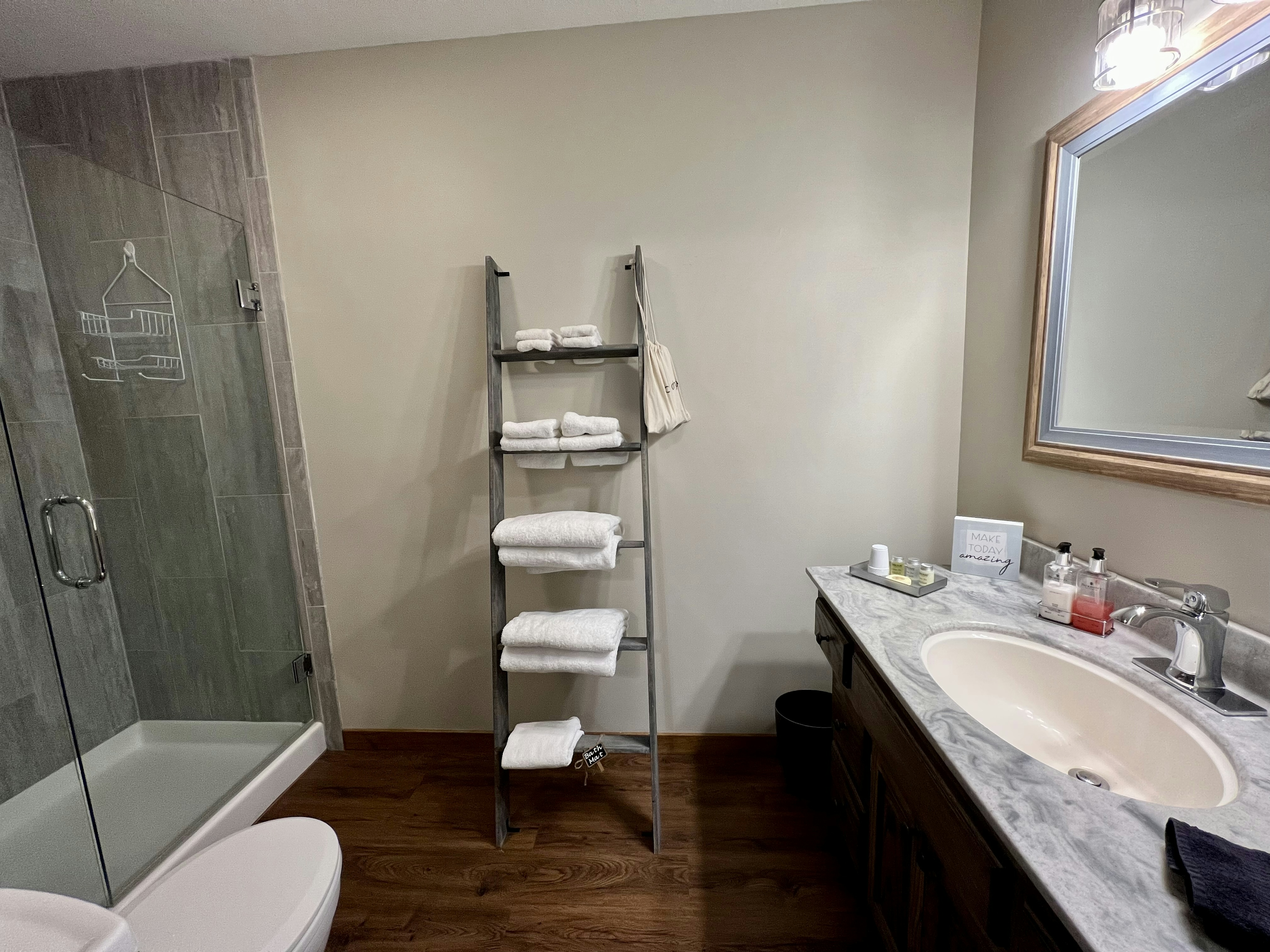 grey tiled shower with glass door, ladder style towel rack with white towels, grey vanity