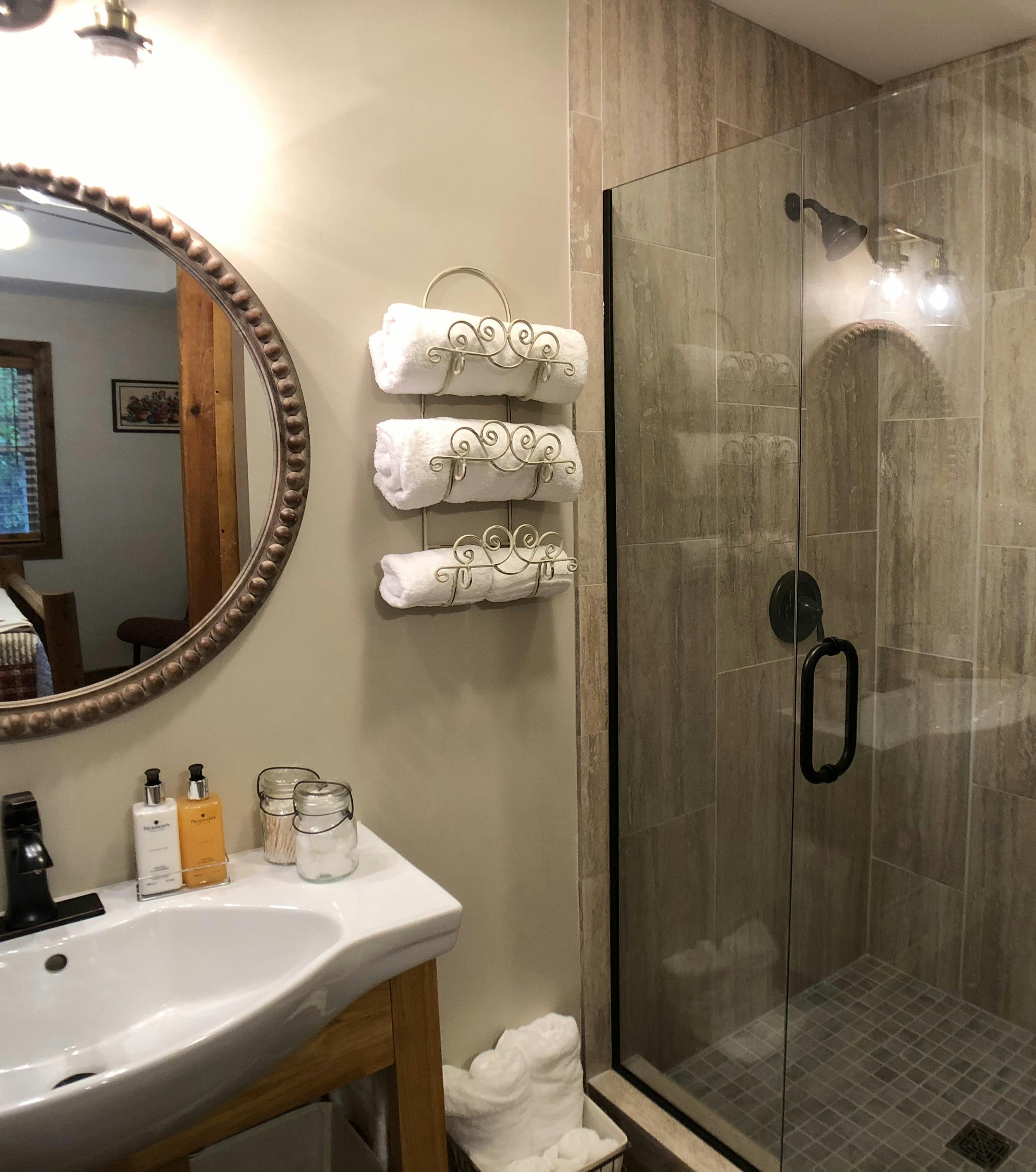 earthtone tiled shower with clear glass door, towel rack on wall with white towels, round mirror above white vanity