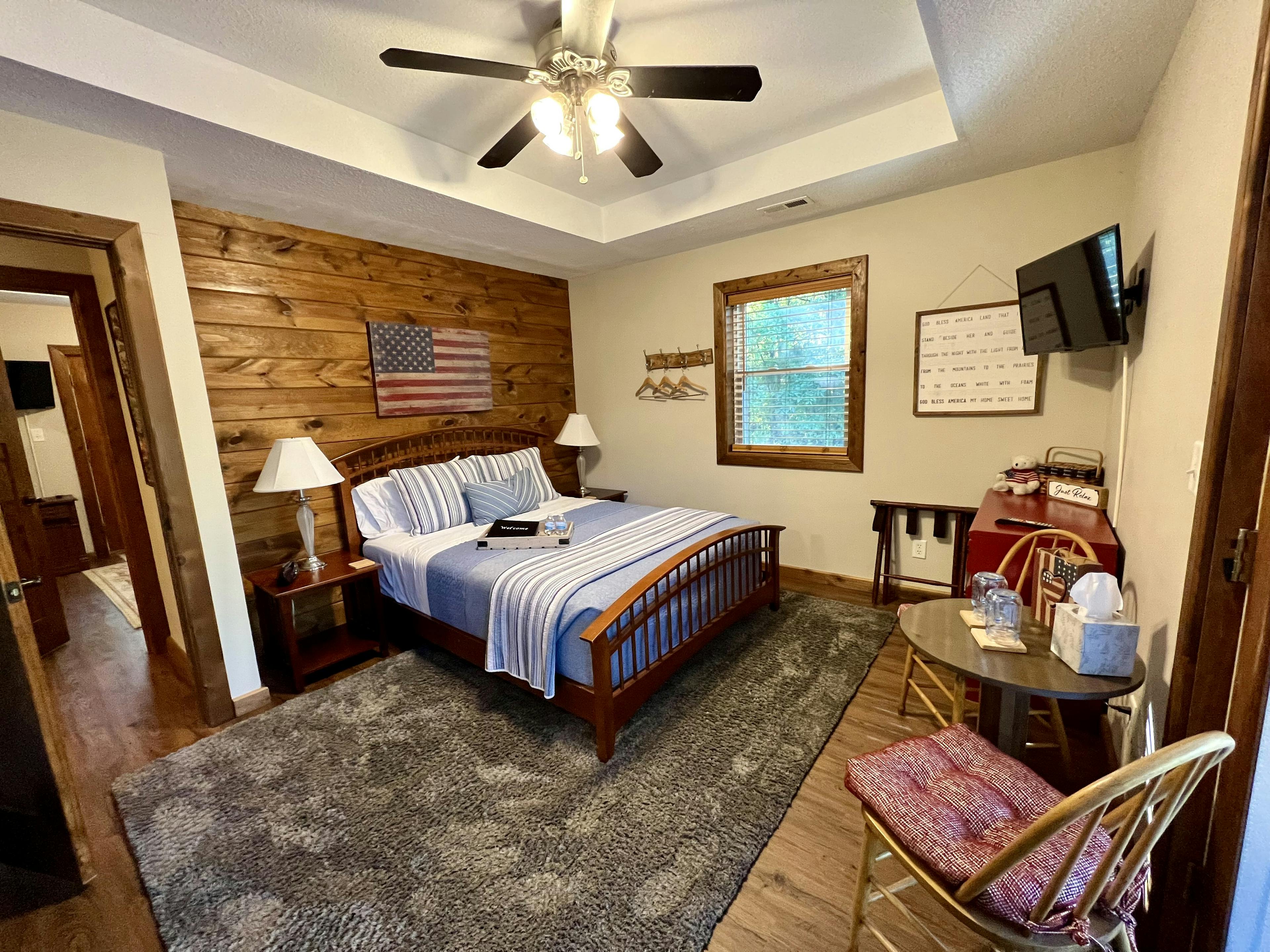 brown wooden bed with light blue quilt, two wooden chairs with pillow, two side tables with lamps, American Flag picture above bed